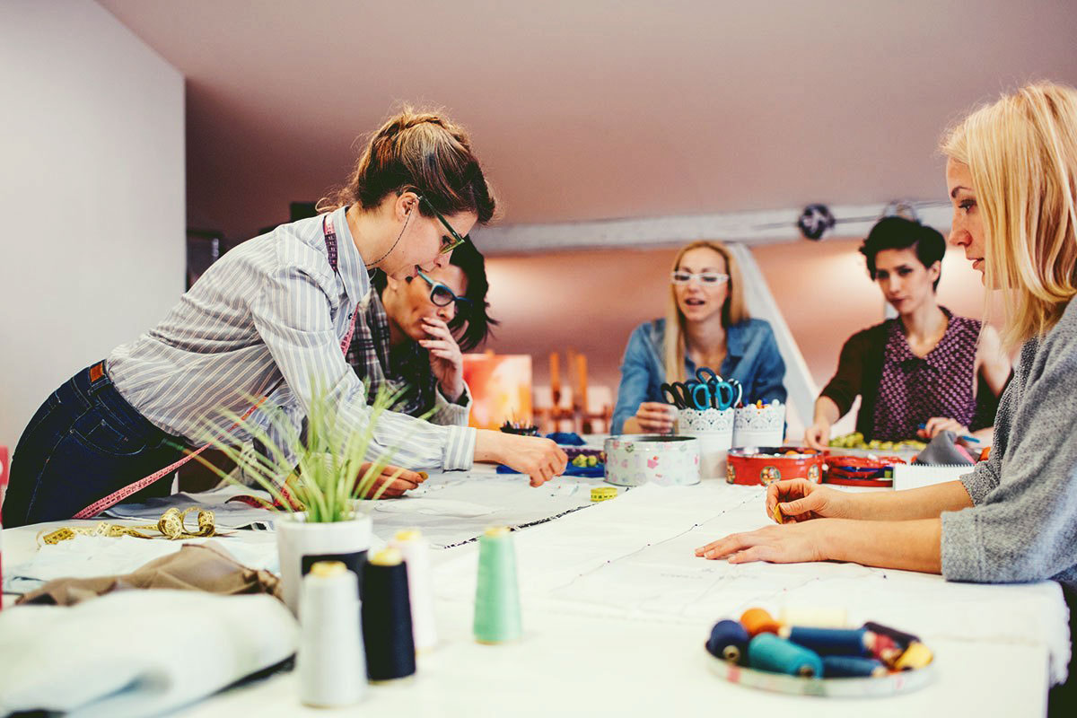 Costura Cinco mulheres reunidas, uma mesa na cor branca, tecido, linhas de costura, fita métrica, conversando e apreendendo técnicas de costura.