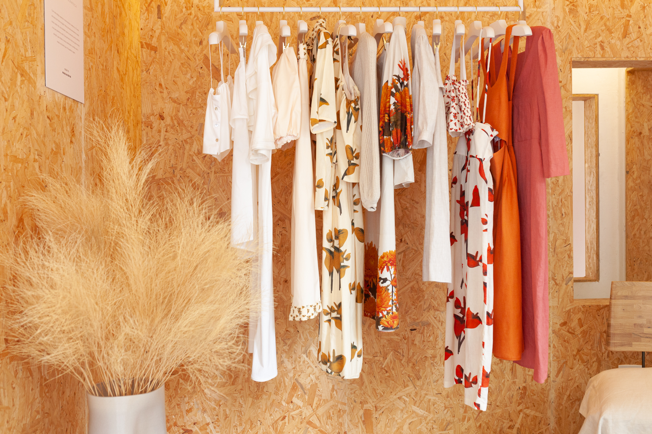 Guarda roupa sustentável Parede rustica, cor caramelo com uma arara no centro com roupas e um vaso de planta do lado esquerdo.