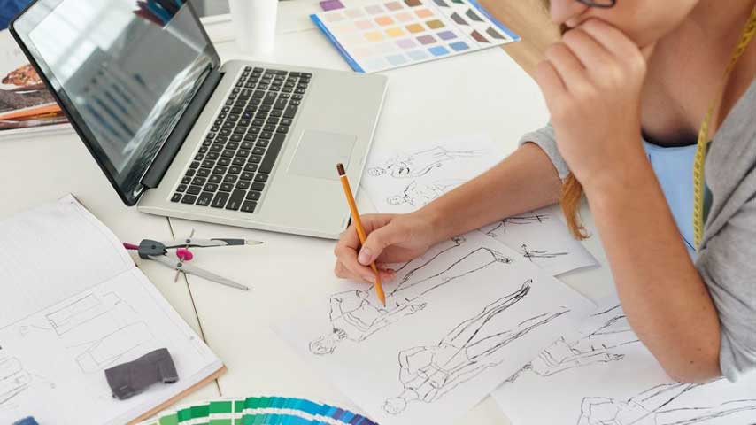 Mulher sentada em frente um notebook e desenhando manualmente croqui de moda.