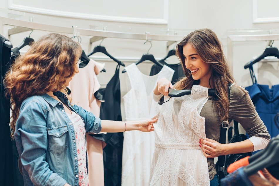 Duas mulheres juntas em uma loja de roupas, sendo uma loira e uma morena, a loira orientando a morena quanto aos detalhes da peça de roupa. 